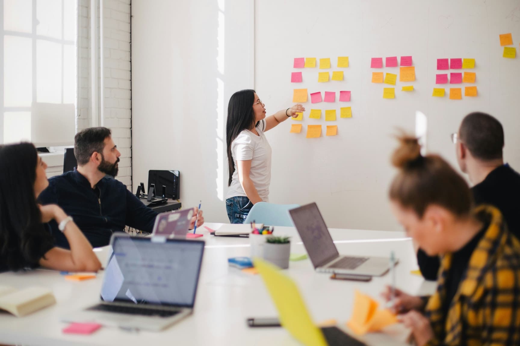 A meeting with several people working on notes, viewing laptops, and discussing things. One woman is up front with sticky notes coordinating the meeting.