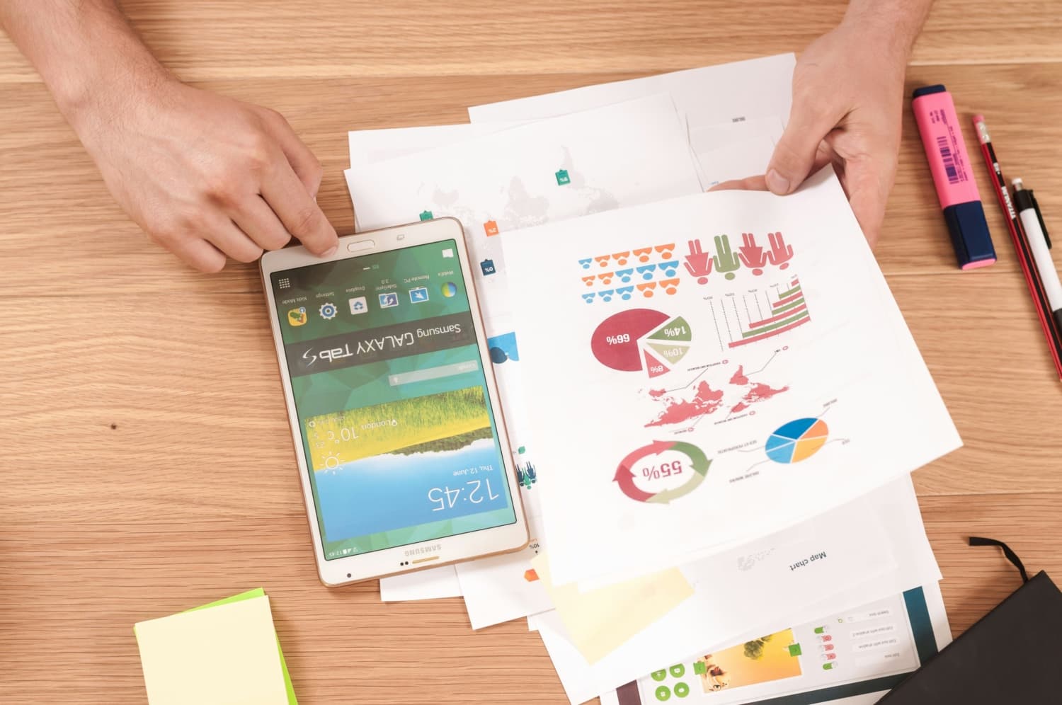 A person looking at sheets of paper detailing user demographics and statistics with a tablet, pen, pencil, highlighter, and notebook nearby.