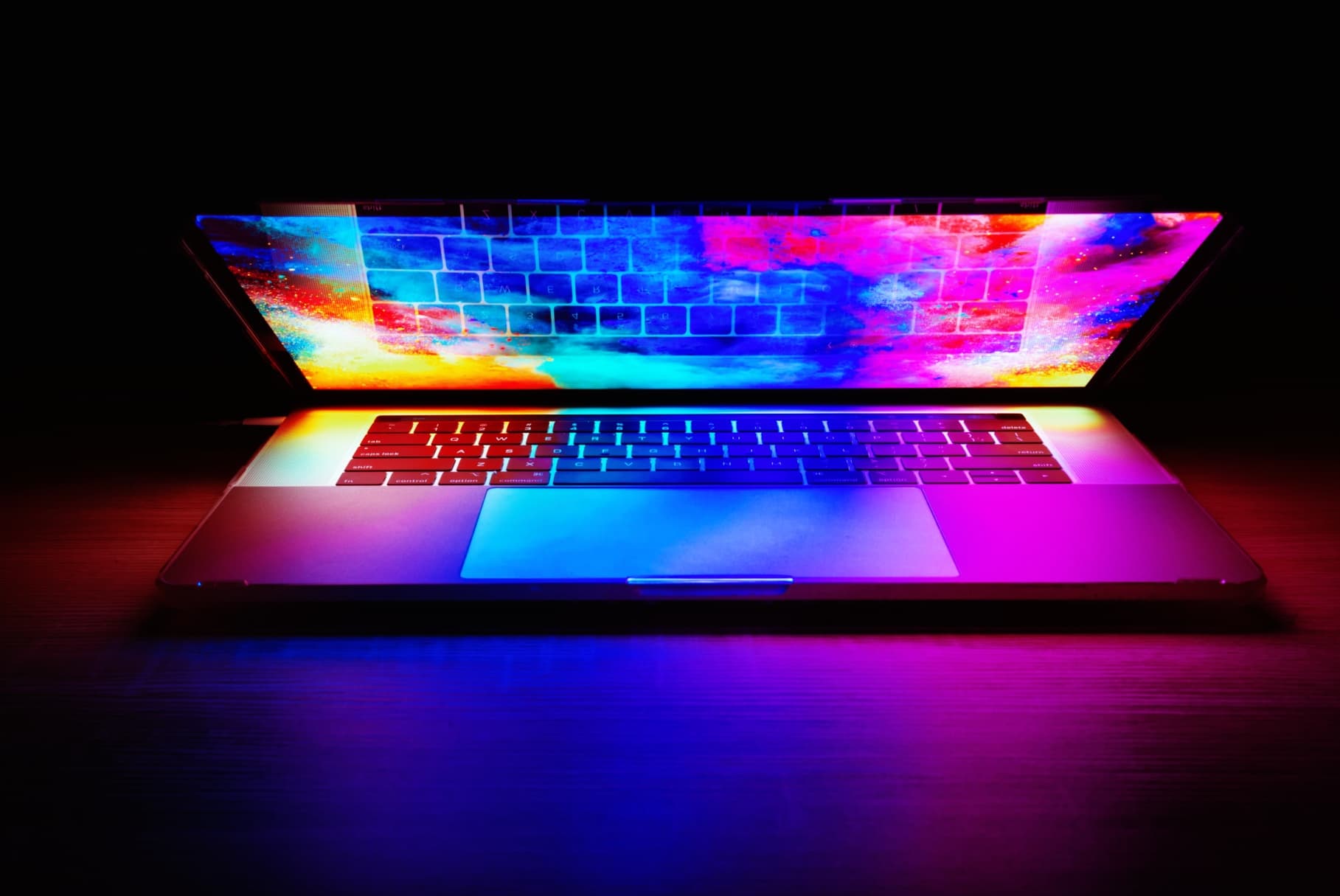 A MacBook Pro in the dark on a wood desk with the lid half shut and a wallpaper of beautiful abstract color that’s casting a light on the keyboard and around the edge of the laptop.