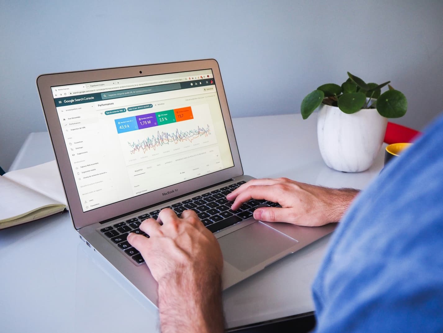 A man's hands on the keyboard of his MacBook Air with the Google Search Console on the screen viewing site analytics, with a coffee mug, plant, and notebook on the desk next to him.