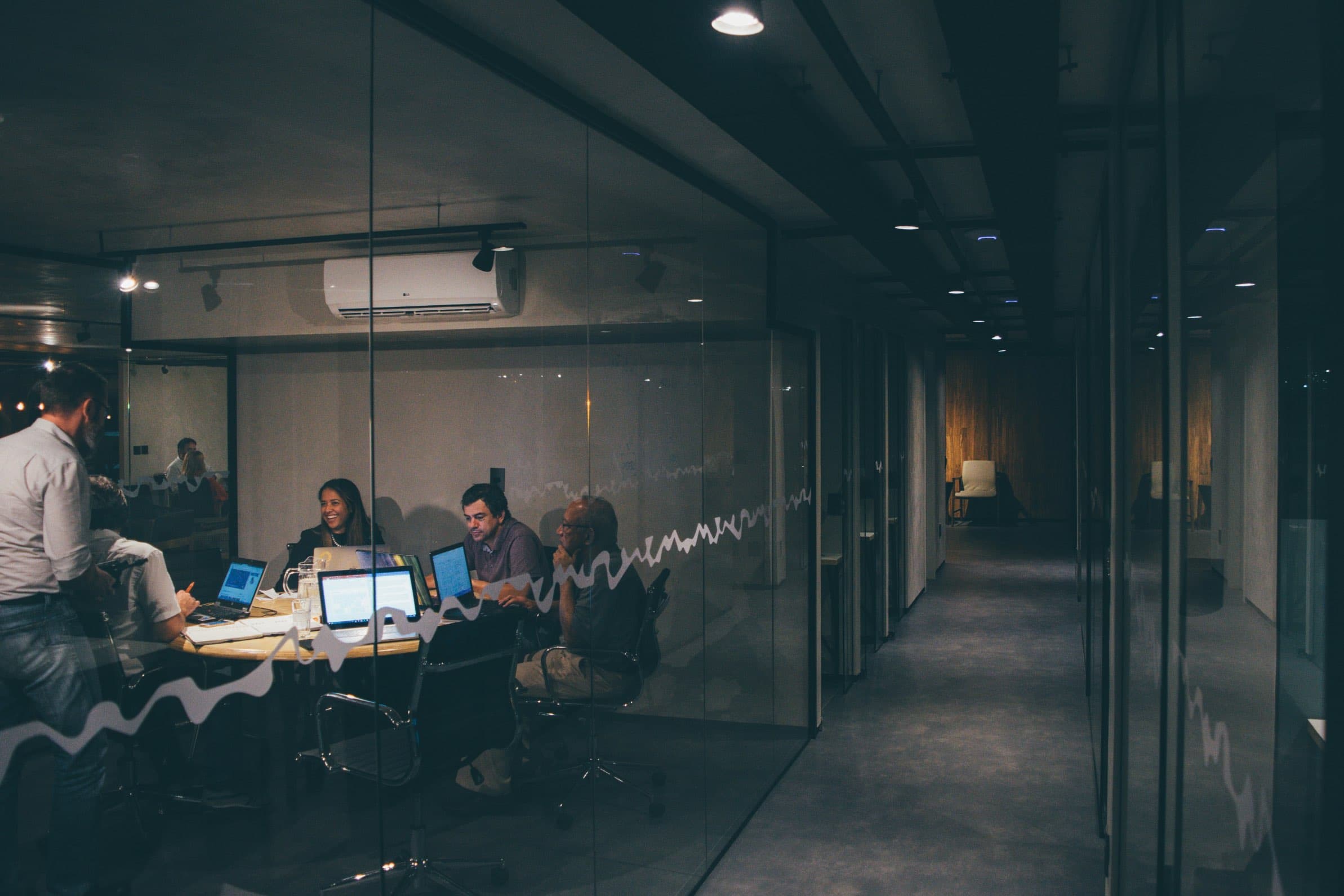 A view of an office space with the hallway on the right lined by glass offices, with a glass conference room on the left through which can be seen a meeting of several people working on their laptops around a table, some smiling while others look deep in thought.