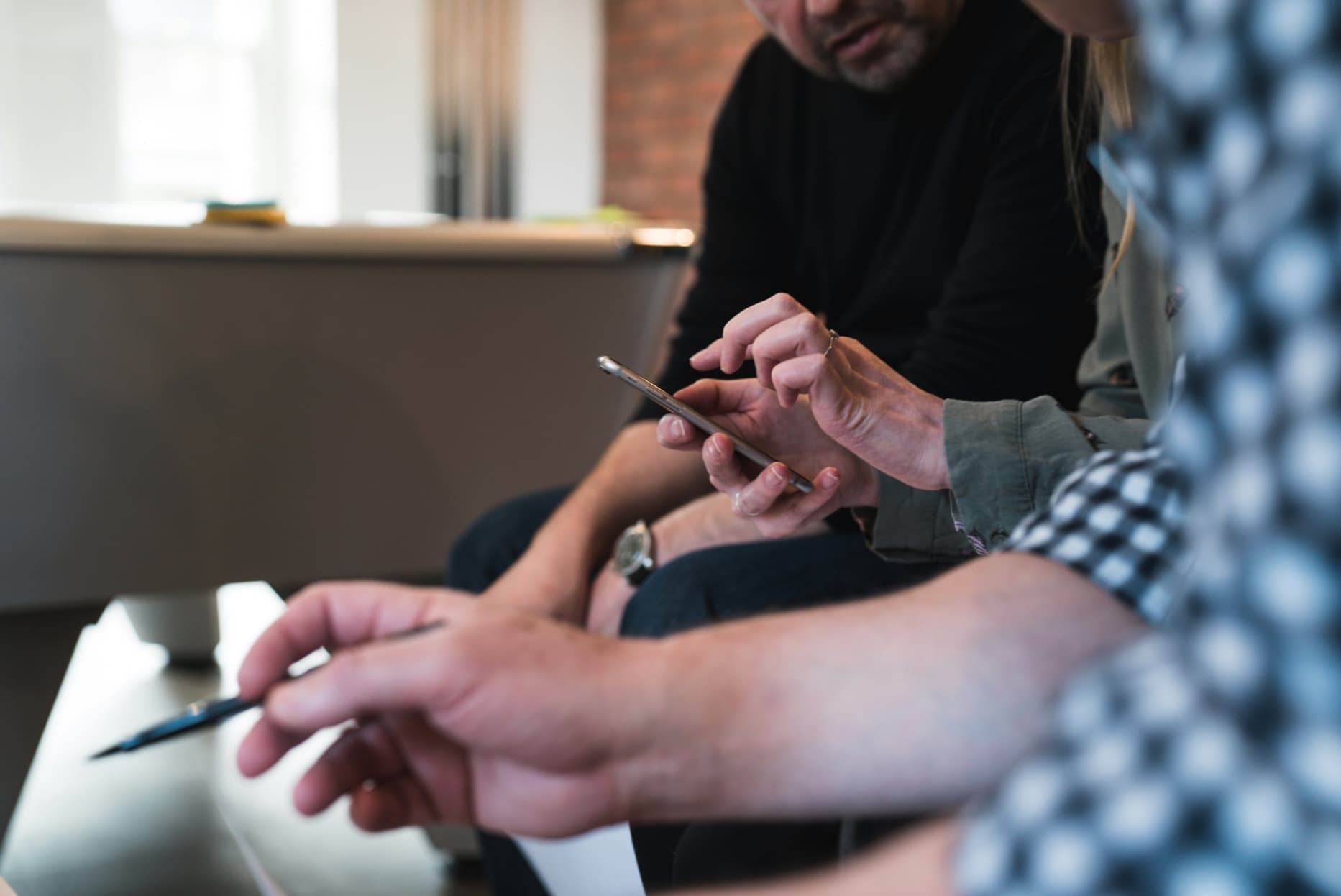 A group of three people discussing and taking notes as they test something on a smartphone.