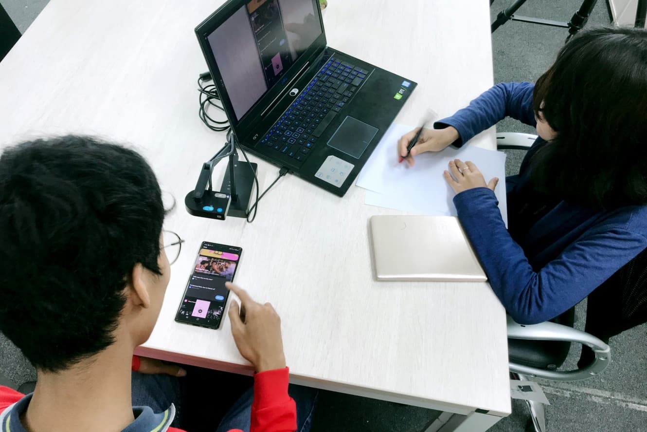 A young man testing a design on his phone next to a young woman taking notes while looking at the working model of that design on her laptop.